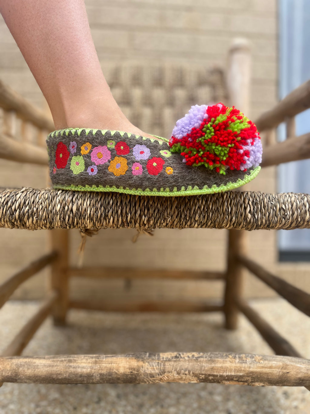 Embroidered woollen slipper with floral pattern and colourful pom-pom on a wooden chair.