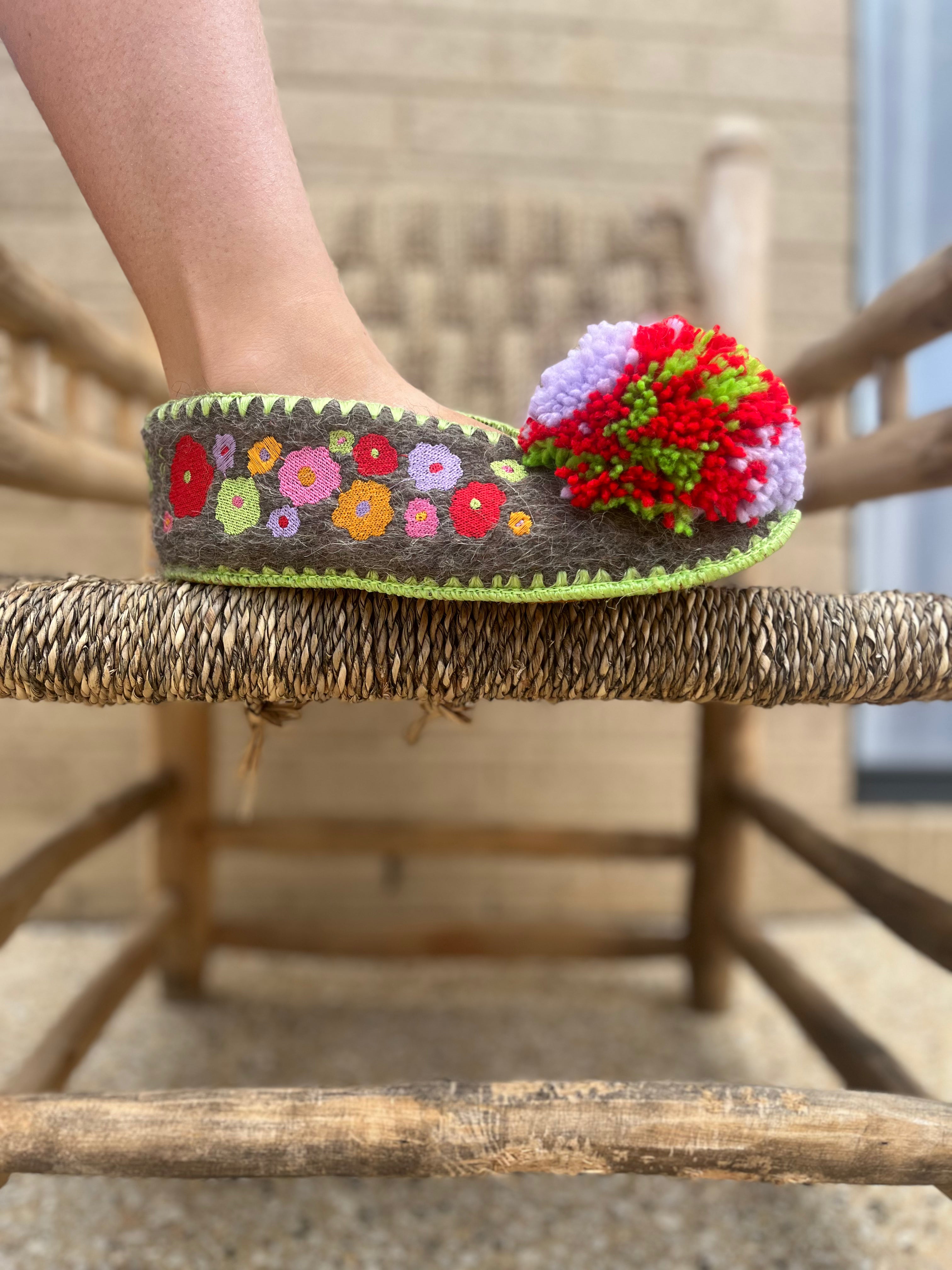 Embroidered woollen slipper with floral pattern and colourful pom-pom on a wooden chair.